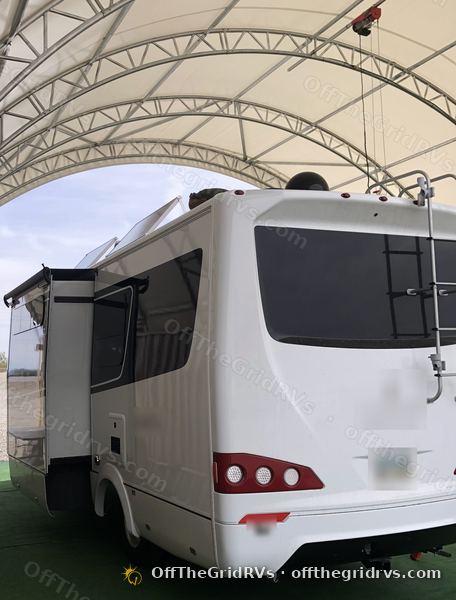Rear view of a white Leisure Travel Vans Unity under the OffTheGridRVs shop canopy, its roof solar panels tilted up on manual tilt brackets